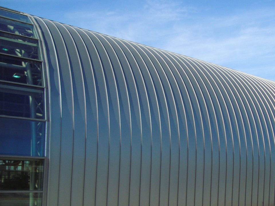 Curved aluminium roof of a large hall in front of a blue sky.