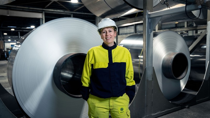 A smiling Speira employee in work clothes in front of stacked aluminum coils.